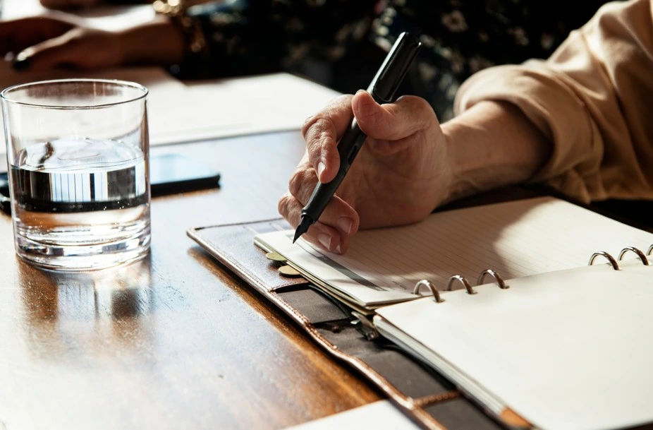 Hand writing in notebook on desk with glass of water