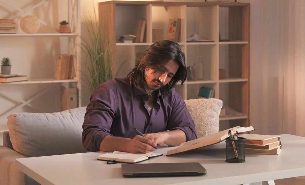 man writing in notebook at desk with books and laptop