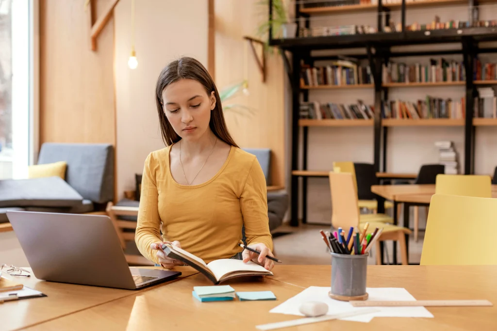 A woman studies in a library, surrounded by books and a laptop. The setting is cozy and inviting with warm lighting.