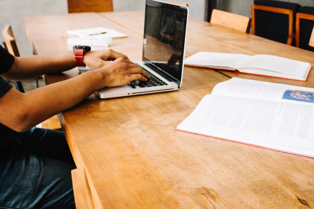 Person typing on a laptop at a table, surrounded by open books and a notebook, creating a focused and studious atmosphere.