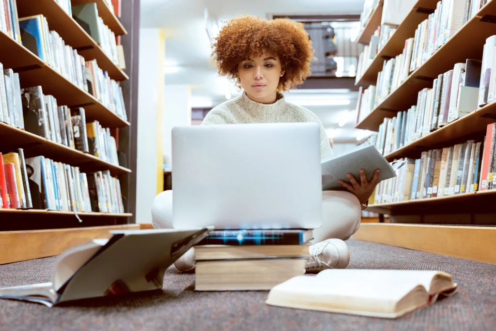 Young woman sits on a library floor, focused on a laptop propped on books. She's surrounded by shelves and appears deep in study.